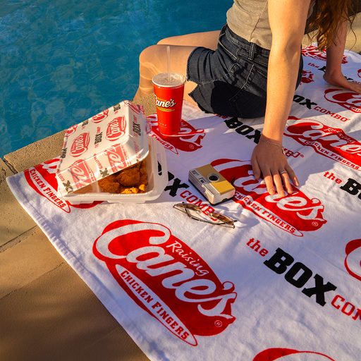 Person sitting on a Raising Cane's Box Combo beach towel by a pool, with food and a drink.