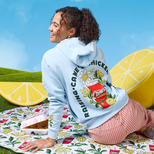Person wearing the Cane’s Fresh Lemon Hoodie with brand logo, sitting on a picnic blanket surrounded by lemons and cake
