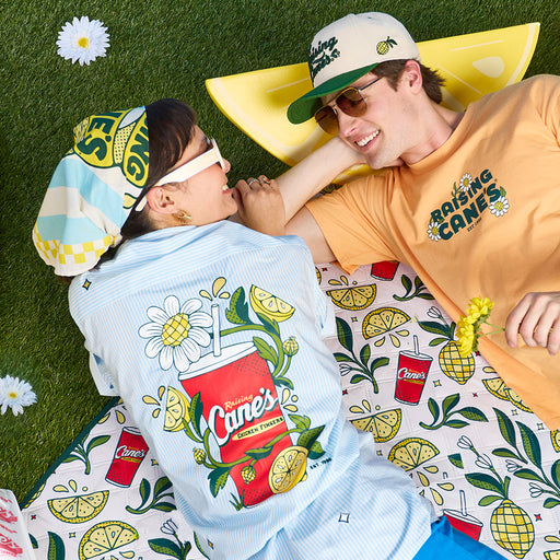 Two people lying on a branded towel and blanket with Cane's' logo on a grassy background