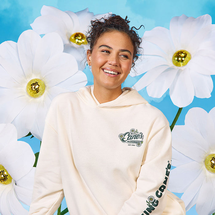 Model wearing the Cane’s Sauce Floral Hoodie, a cream hoodie with a brand logo, standing against a blue background with large white flowers