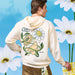 Model wearing the Cane’s Sauce Floral Hoodie a cream hoodie with graphic design standing against a blue sky with white flowers
