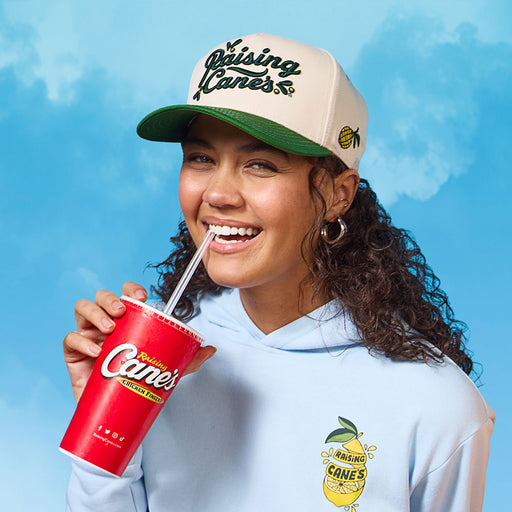 Woman wearing the Cane’s Fresh Lemon Hat a Raising Cane’s cap and holding a red cup with a straw against a blue sky background